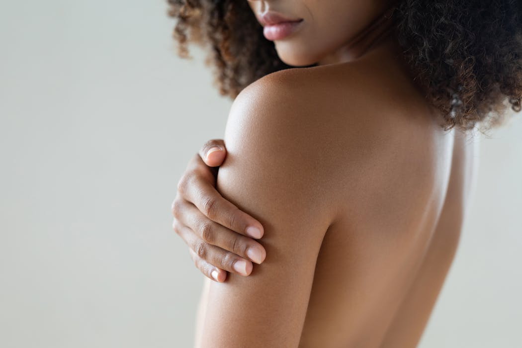 Femme de profil touchant son épaule nue, sur fond neutre.