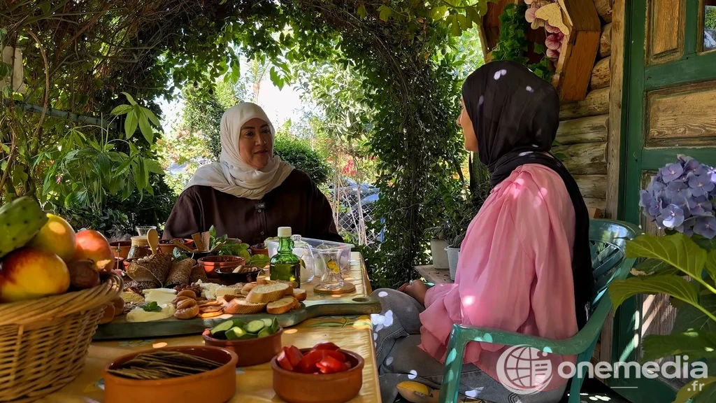 Deux femmes sont assises à une table garnie de produits alimentaires sous une végétation dense.