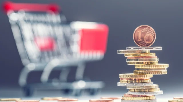 Une pile de pièces surmontée d’une pièce d’un centime, devant un petit chariot de courses flou à poignée rouge.