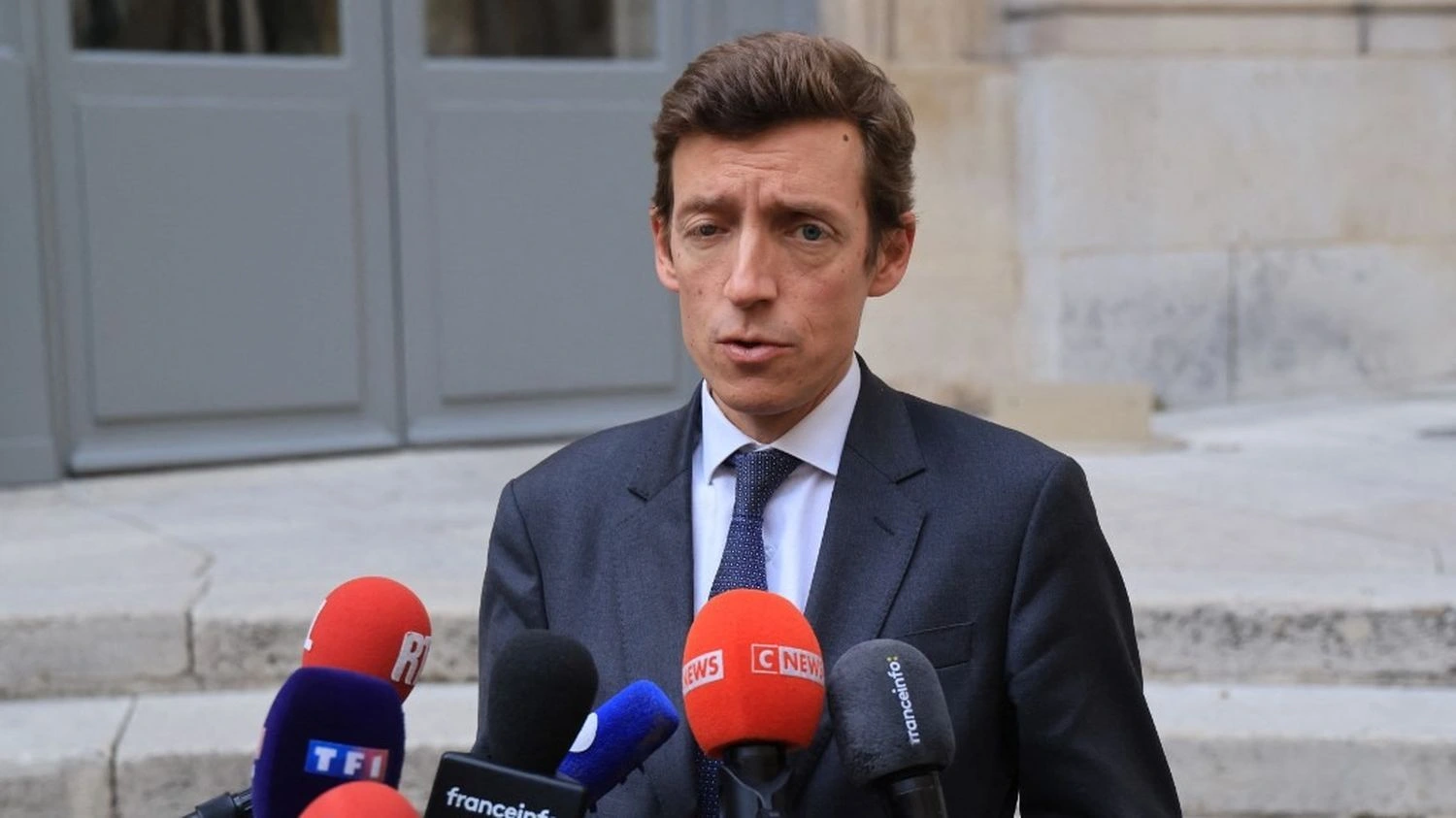 Édouard Geffray, en costume et cravate, s’exprime devant plusieurs microphones à l’extérieur d’un bâtiment.