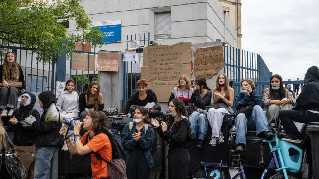 Des jeunes sont réunis devant les grilles d’un lycée avec des pancartes en carton ; une personne au premier plan tient un mégaphone.