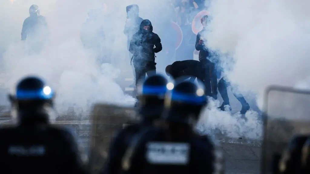 Des policiers casqués vus de dos font face à des personnes partiellement enveloppées de fumée dans une rue.