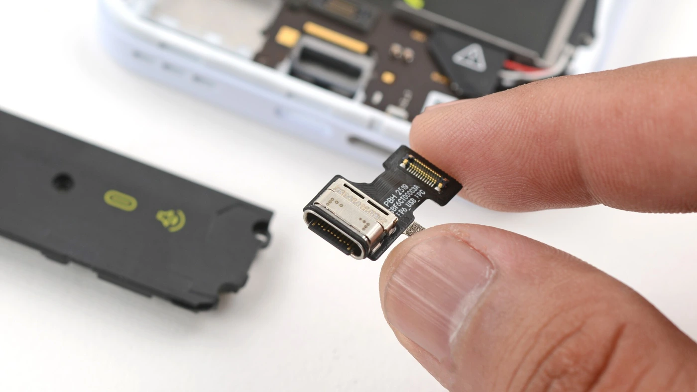 Module USB-C du Fairphone 6 tenu entre deux doigts, avec le téléphone ouvert en arrière-plan.