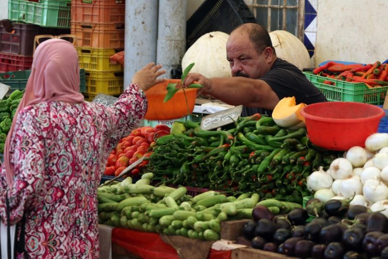 Une femme en foulard rose devant un vendeur, avec des tomates, des poivrons, des oignons et des aubergines sur l’étal.