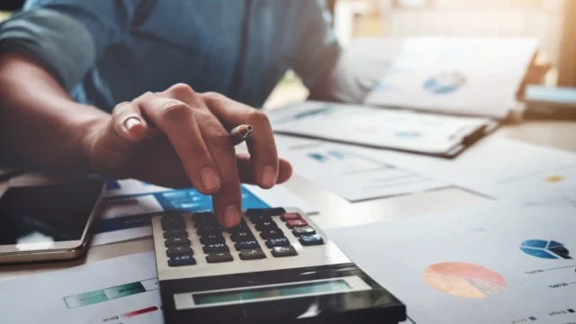 Une personne tenant un stylo utilise une calculatrice sur un bureau couvert de documents avec des graphiques, à côté d’un appareil mobile.